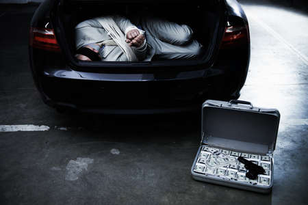 Money through violence. A bound and blindfolded businessman lying in the trunk of a car next to a briefcase of ransom money.の写真素材
