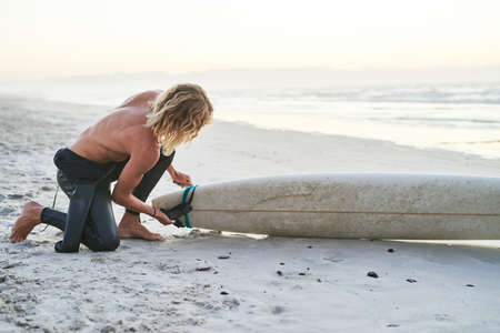 Preparing for a great day in the raging ocean. Full length shot of a handsome young surfer getting his surfboard ready before going surfing at the beach.の写真素材