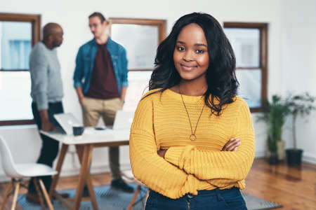 Love the career youre in. Portrait of a cheerful young businesswoman standing with her arms folded and her work colleagues in the background inside of the office.の写真素材