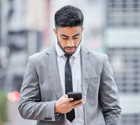 You have until the end of the business day. Shot of a young businessman using his smartphone while walking outside.の写真素材