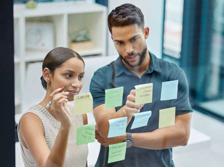 Lets figure this out.... Shot of two businesspeople brainstorming with sticky notes on a transparent...