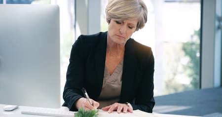Its a busy day in the office. Shot of a mature businesswoman writing notes while working on a computer in an office.の写真素材