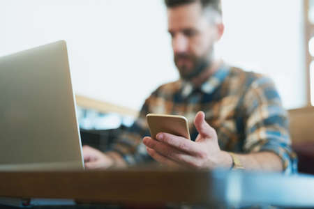 Take your wireless devices wherever you go. Shot of a young man using his cellphone and laptop while sitting in a cafe.の写真素材