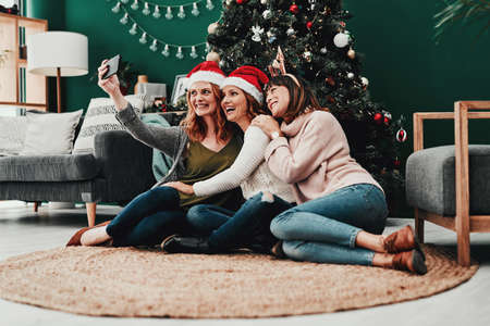 This one is going to the album. Shot of three attractive middle aged women taking self portraits together with a cellphone at home during Christmas time.の写真素材
