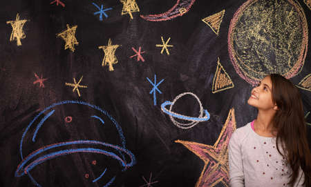 Let your imagination take you anywhere. Shot of a little girl standing in front of a chalkboard drawing.の写真素材
