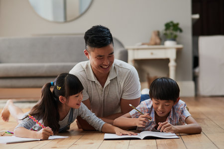 Spending time with my children. Shot of a cheerful father and his two children doing homework together while lying on the floor at home during the day.の写真素材