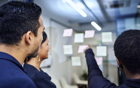 Lets come up with a master plan together. Rearview shot of a group of businesspeople brainstorming with notes on a glass wall in an office.の写真素材