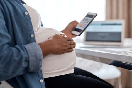 This app has helped me so much through my pregnancy journey. Cropped shot of a pregnant woman using her cellphone while sitting at home.の写真素材