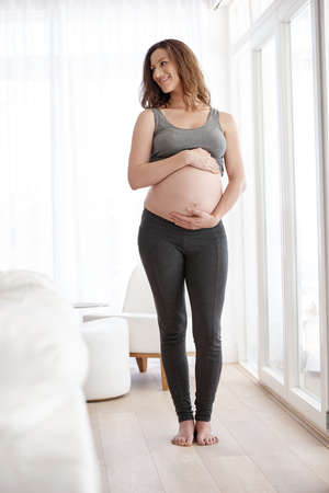 Not long now.... Full length shot of a young pregnant woman standing in her home.