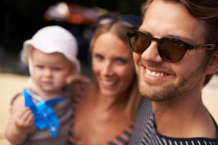 Hes a dashing dad. A a smiling handsome man standing with wife and baby in the background.