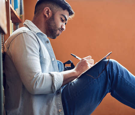 An investment in knowledge pays the biggest dividends. Shot of a young man making notes in a library at university.の写真素材