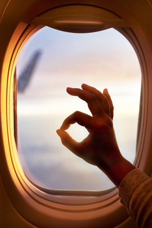 This flight is going just fine. Shot of a passenger making an okay sign while looking through the window of an airplane.の写真素材