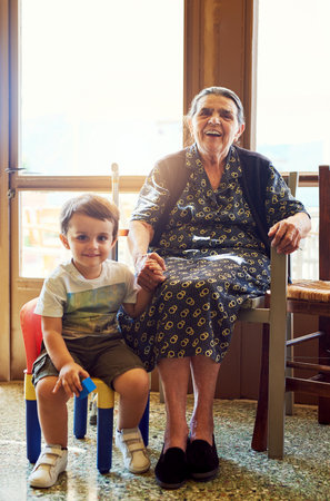 The youth and the wise. Portrait of a cheerful little boy sitting next his great grandmother while looking into the camera at home.の写真素材