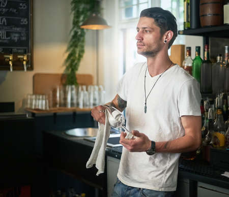 Daydreaming on duty. Shot of a young waiter daydreaming while polishing a glass behind the bar.の写真素材