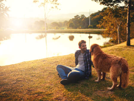You having fun. Full length shot of a handsome young man and his dog spending a day by a lake in the park.の写真素材