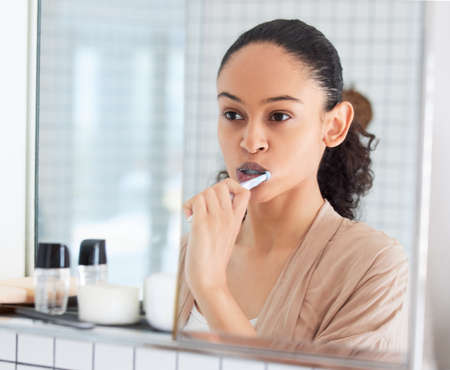 Attending to those pearly whites. Shot of an attractive young woman standing alone in her bathroom at home and brushing her teeth in the morning.の写真素材