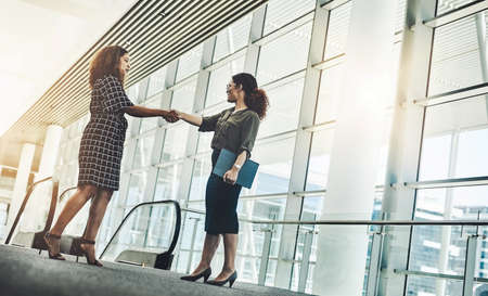 Two go getters coming together. Low angle shot of two attractive young businesswomen shaking hands while standing in a modern workplace.の写真素材