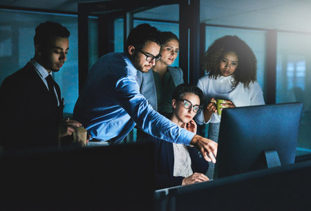 Its taking longer than we thought. Shot of colleagues standing together as they work on something on a computer at night.の写真素材