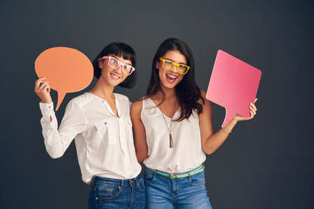 We always think the same. Studio shot of two cheerful young women holding up speech bubbles while standing against a dark background.の写真素材