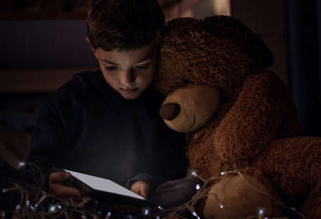 The kingdom belongs to little children. Shot of an adorable little boy using a digital tablet with his teddy bear at night in his bedroom.の写真素材