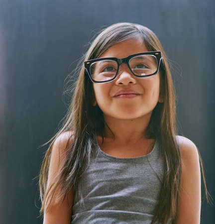 Shes always at wonder about the world. Studio shot of a little girl wearing spectacles against a dark background.の写真素材