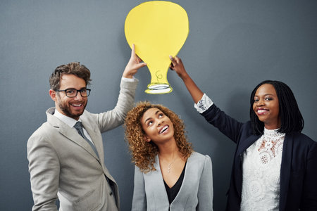 She always comes up with a bright idea. Studio portrait of three businesspeople standing against a gray background with a cutout lightbulb above their head.の写真素材