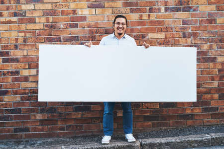 What would you do with all of this space. Shot of a young man holding a blank banner against an urban background outdoors.の写真素材