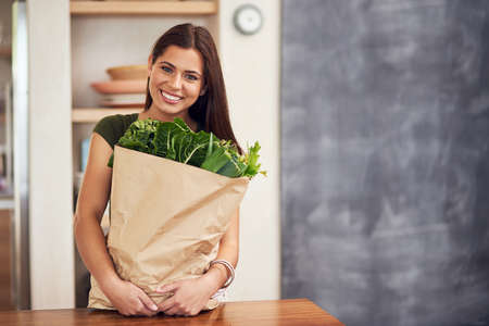 As fresh as it gets. Portrait of an happy young woman holding a bag of groceries in her kitchen.の写真素材