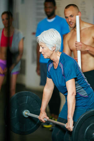 Nothing is going to happen in your comfort zone. Shot of a senior woman lifting weights while a group of people in the background watch on.の写真素材