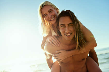 Beach days are a must in summer. Portrait of a young man giving his girlfriend a piggyback ride at the beach.の写真素材