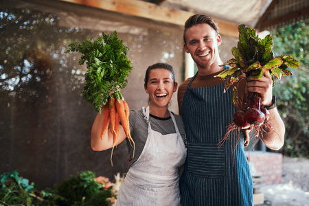 Pick your favorite. Portrait of a happy young couple posing together holding bunches of freshly picked carrots and beetroot at their farm.の写真素材