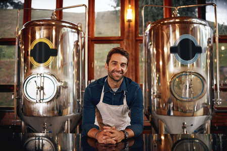 If you want beer we have it. Portrait of a cheerful young barman leaning on the bar counter waiting for customers to serve inside of a beer brewery.の写真素材