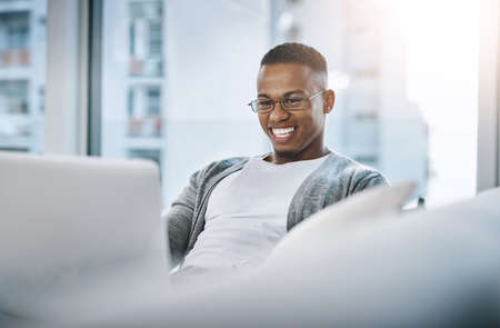 This is what I actually do on my off days. Shot of a handsome young man using his laptop while sitting on a sofa at home.の写真素材
