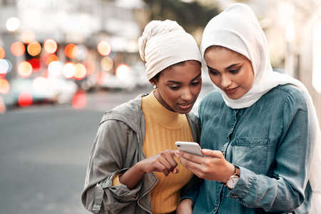 Check the maps. Cropped shot of two attractive young women wearing headscarves and standing together while using a cellphone in the city.の写真素材