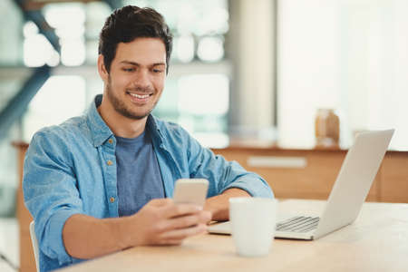 Always connected. Shot of a handsome young man using his cellphone and laptop at home.