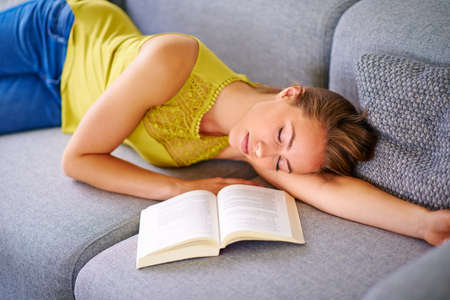 Nowhere more relaxing than home.... Shot of a young woman sleeping beside a book on her sofa at home.の写真素材