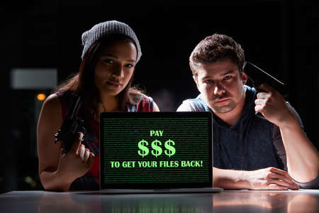Give us the money and no one gets hurt. Shot of two armed hackers using a laptop with Pay $$$ to get your files back on it.の写真素材
