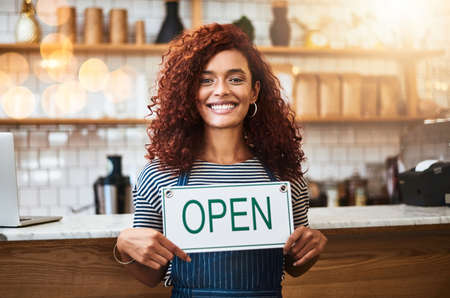 Service with a big smile. Portrait of a young woman holding up an open sign in her store.の写真素材