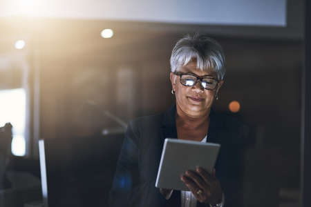 Digital business dealings after dark. Shot of a mature businesswoman using a digital tablet late at night in a modern office.の写真素材