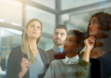 Trying to see their problems as opportunities. Shot of a group of young entrepreneurs brainstorming with notes on a glass wall in an office.の写真素材