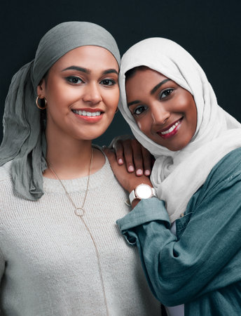 Friends until the end. Cropped shot of two attractive young women wearing hijabs and standing close together against a black background in the studio.の写真素材