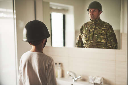 Daddy, come home. Rearview shot of a young boy wearing an army helmet seeing his fathers reflection in the mirror.の写真素材