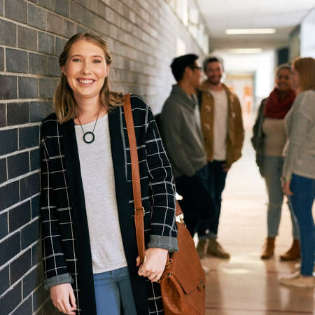 Im enjoying my first year on campus. Portrait of a young female university student standing in a campus corridor with her classmates in the background.の写真素材