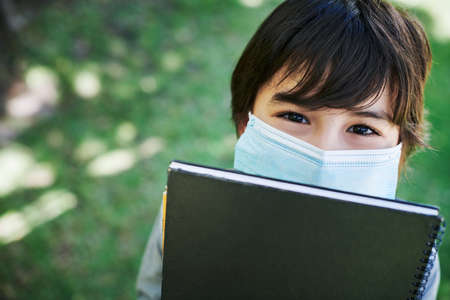 Two things you should never forget. Shot of a little boy wearing a mask and holding books in nature.の写真素材