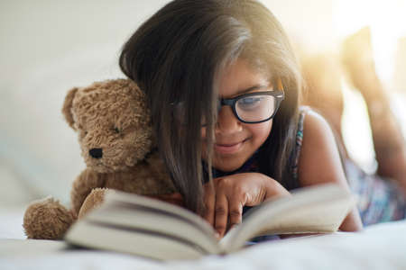 We love storytime, anytime. Shot of a cute little girl reading a book in her bedroom with her teddybear by her side.の写真素材