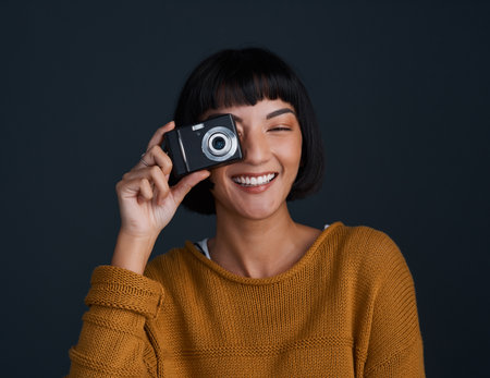 Smile, youre on camera. Studio portrait of a young woman using a camera against a dark background.の写真素材