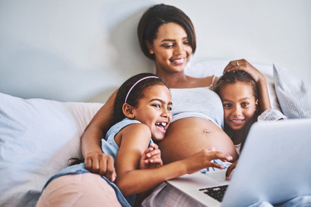 Story time with the family. Shot of two cheerful little girls relaxing next to their pregnant mother while watching a movie on a laptop at home during the day.の写真素材