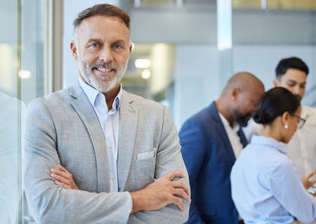 Leading a thriving empire. Portrait of a mature businessman standing in an office with his colleagues in the background.の写真素材