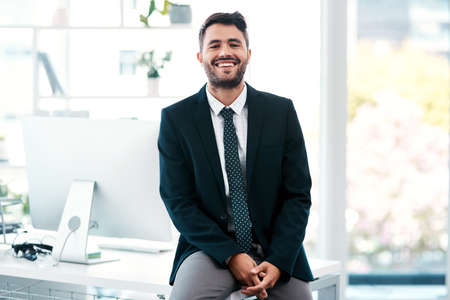 Id like to think the white collar life chose me. Cropped portrait of a handsome young businessman smiling while sitting on his desk in a modern office.の写真素材