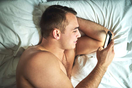 Being intimate online. Cropped shot of a young handsome shirtless man using a cellphone in bed at...